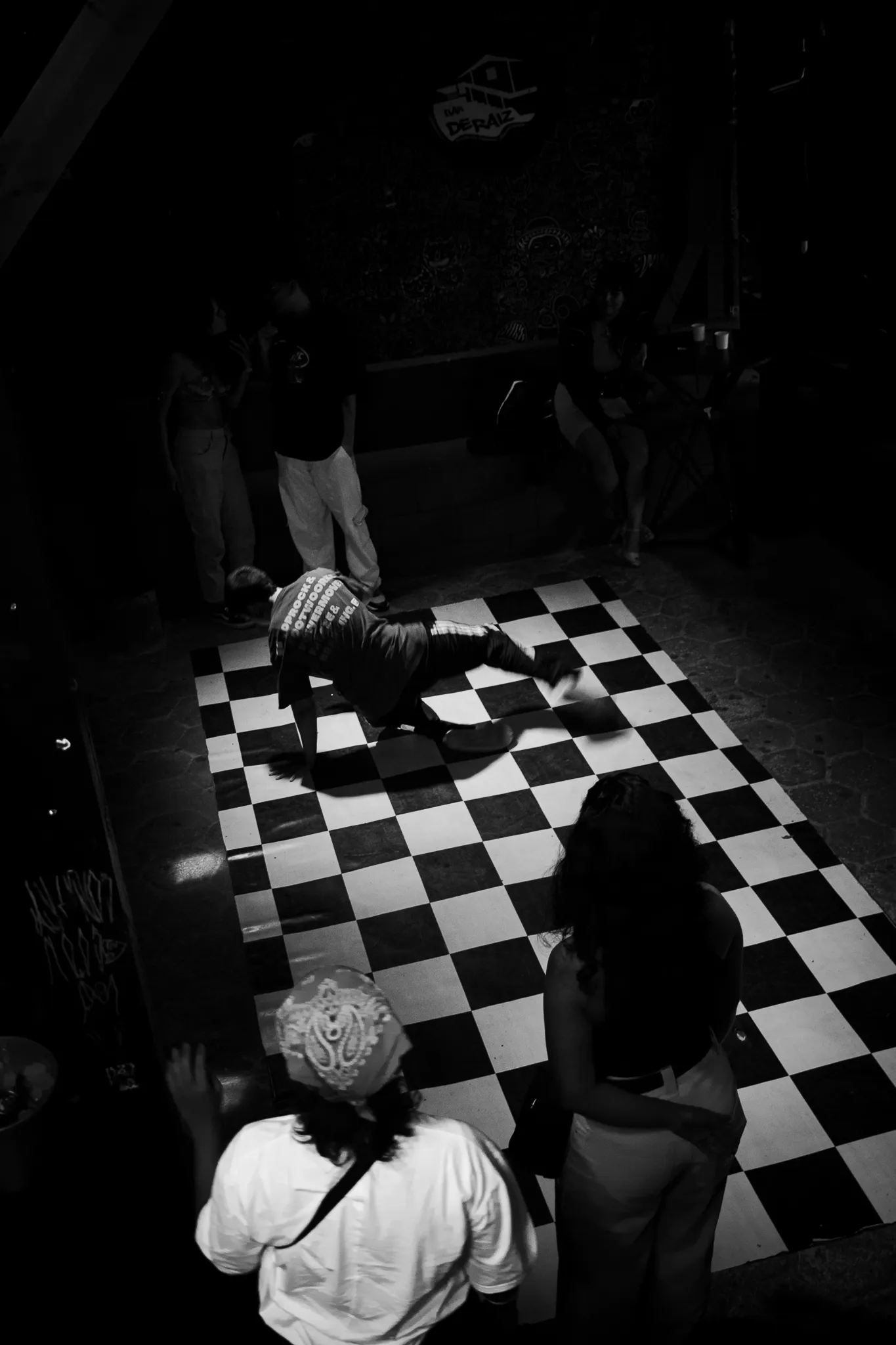 Foto em preto e branco, vista de cima: um b-boy no chão sobre um tapete xadrez, no meio da roda, com gente em volta assistindo.
